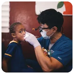 Dr. Lee wearing magnifying loupes and a mask examining a young child's mouth during a dental mission trip